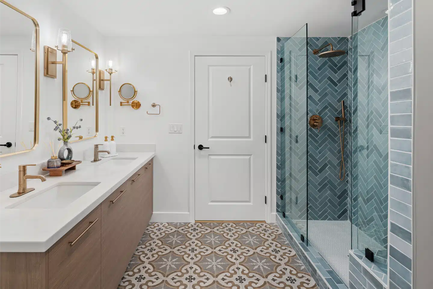 Primary bathroom with a blue-green herringbone tile shower, floating oak vanity, brass fixtures and patterned floor tile