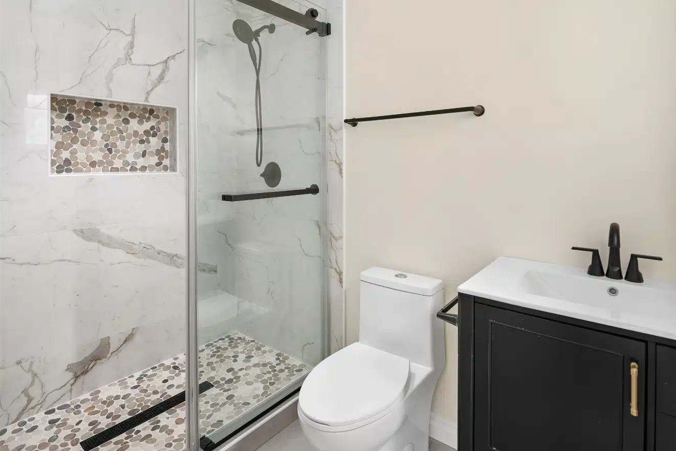 The rebuilt bathroom with a frameless glass shower, dark vanity and pebble mosaic band