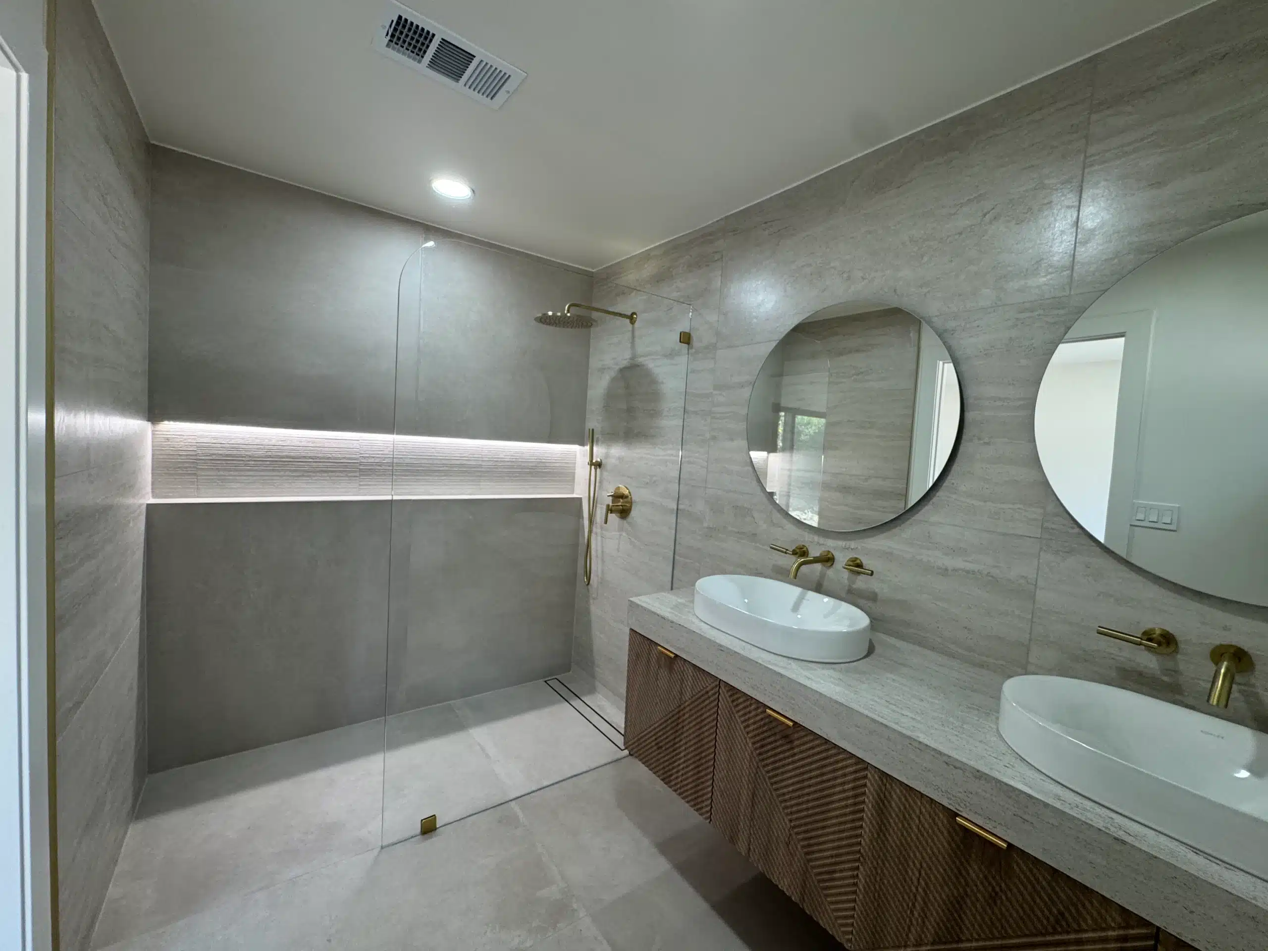 Primary bathroom with a floating oak vanity, twin round mirrors, brass fixtures and a backlit niche in large-format stone-look porcelain