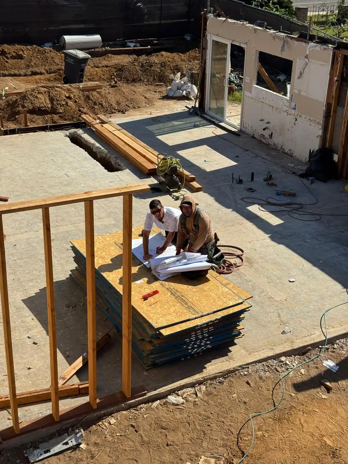 Two people reviewing construction plans beside exposed wall framing and stacked wood panels