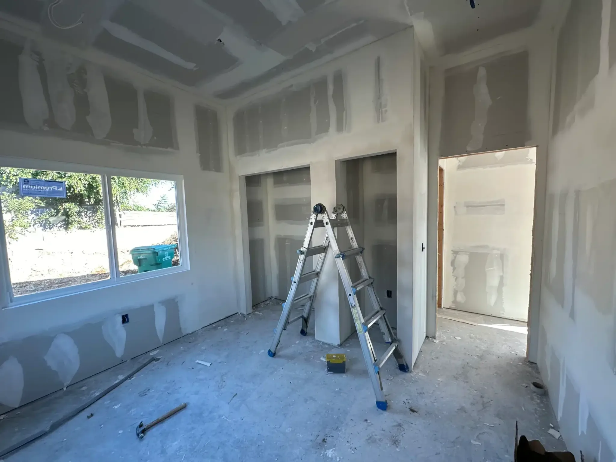 Unfinished interior room with white drywall walls, metal ladder, and windows during construction.