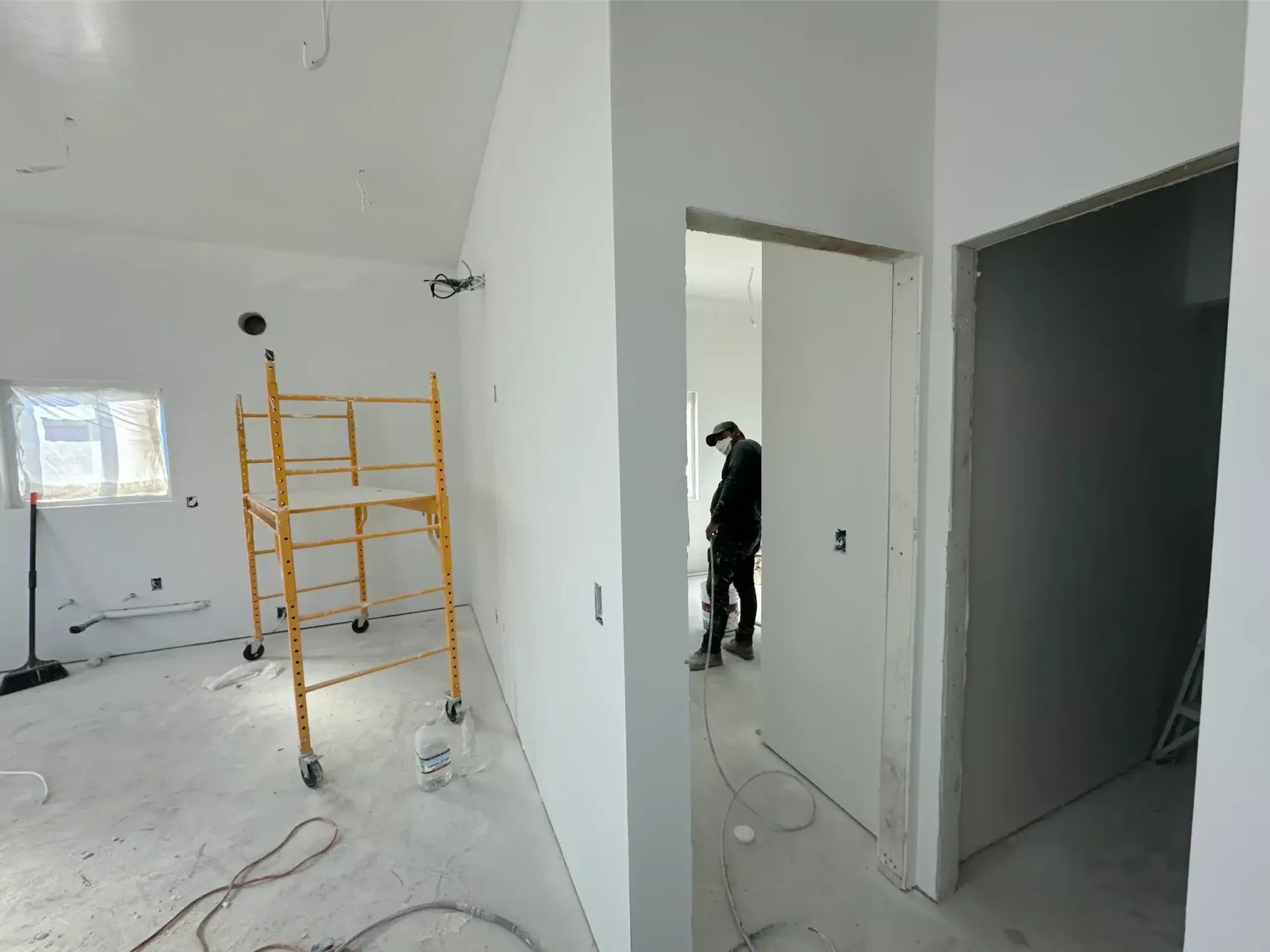Unfinished room with white drywall walls, yellow metal scaffolding, and a worker in another room through an open doorway.