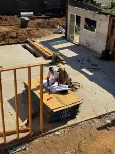 Two workers review blueprints on stacked panels at an active construction site with exposed wooden framing.